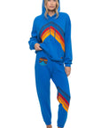 A woman stands barefoot in Aviator Nation's all seasons circle chevron women's sweatpant, a comfy vintage-inspired set with colorful rainbow stripes and a matching patch on the waistband. One hand is in her pocket, the other holding her hoodie.
