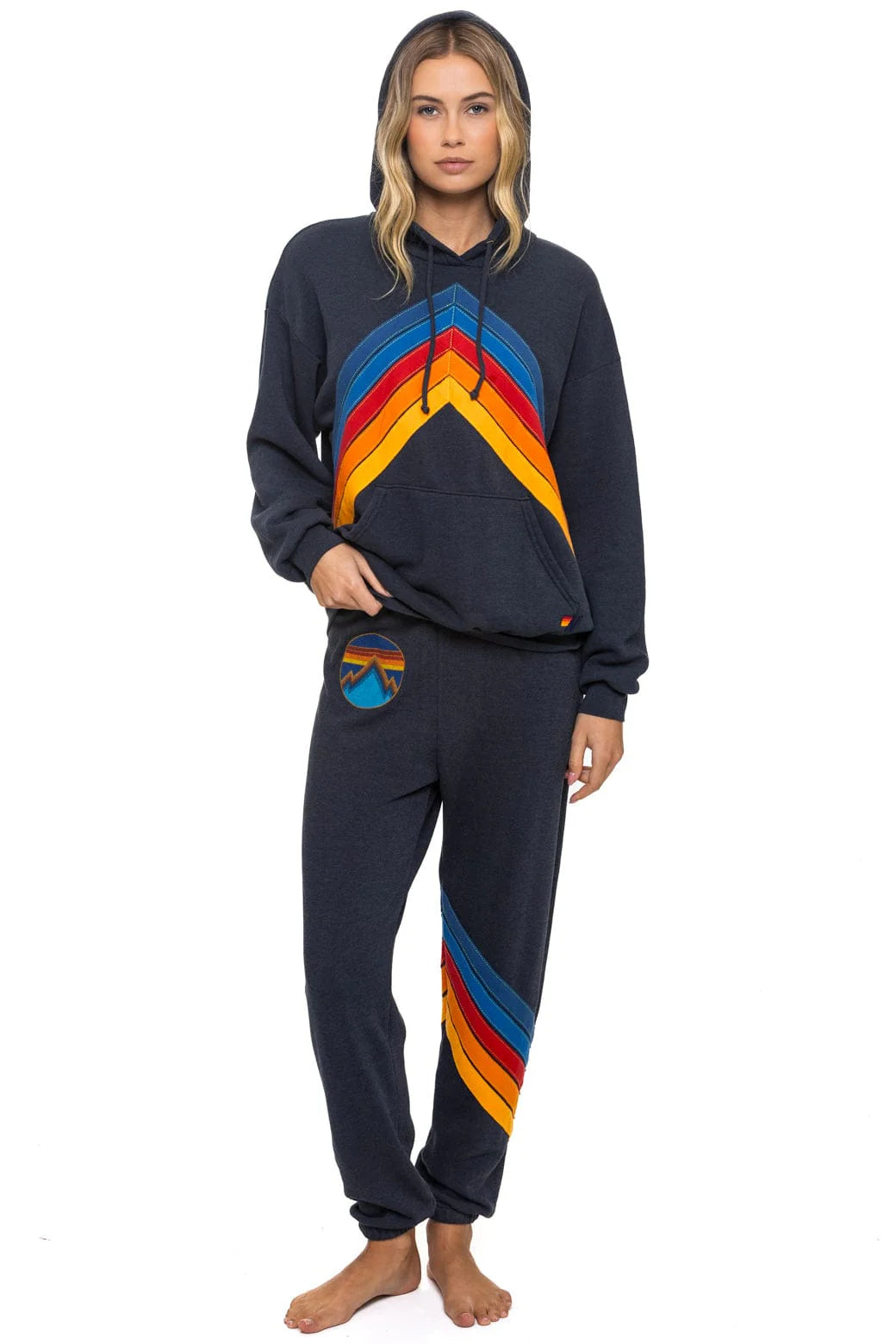 A woman stands barefoot in Aviator Nation’s all seasons circle chevron women's sweatpant, paired with a dark hoodie featuring colorful stripes and vintage vibes. Wavy blonde hair and a neutral expression complement the hand-sewn Los Angeles design.