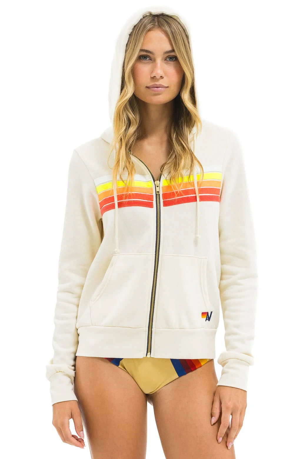 A woman models the Aviator Nation 5 stripe - zip hoodie with bold red, orange, and yellow stripes across the chest, hood up, paired with a tan bikini bottom with colorful stripes—showcasing the retro-inspired style.
