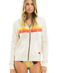 A woman models the Aviator Nation 5 stripe - zip hoodie with bold red, orange, and yellow stripes across the chest, hood up, paired with a tan bikini bottom with colorful stripes—showcasing the retro-inspired style.