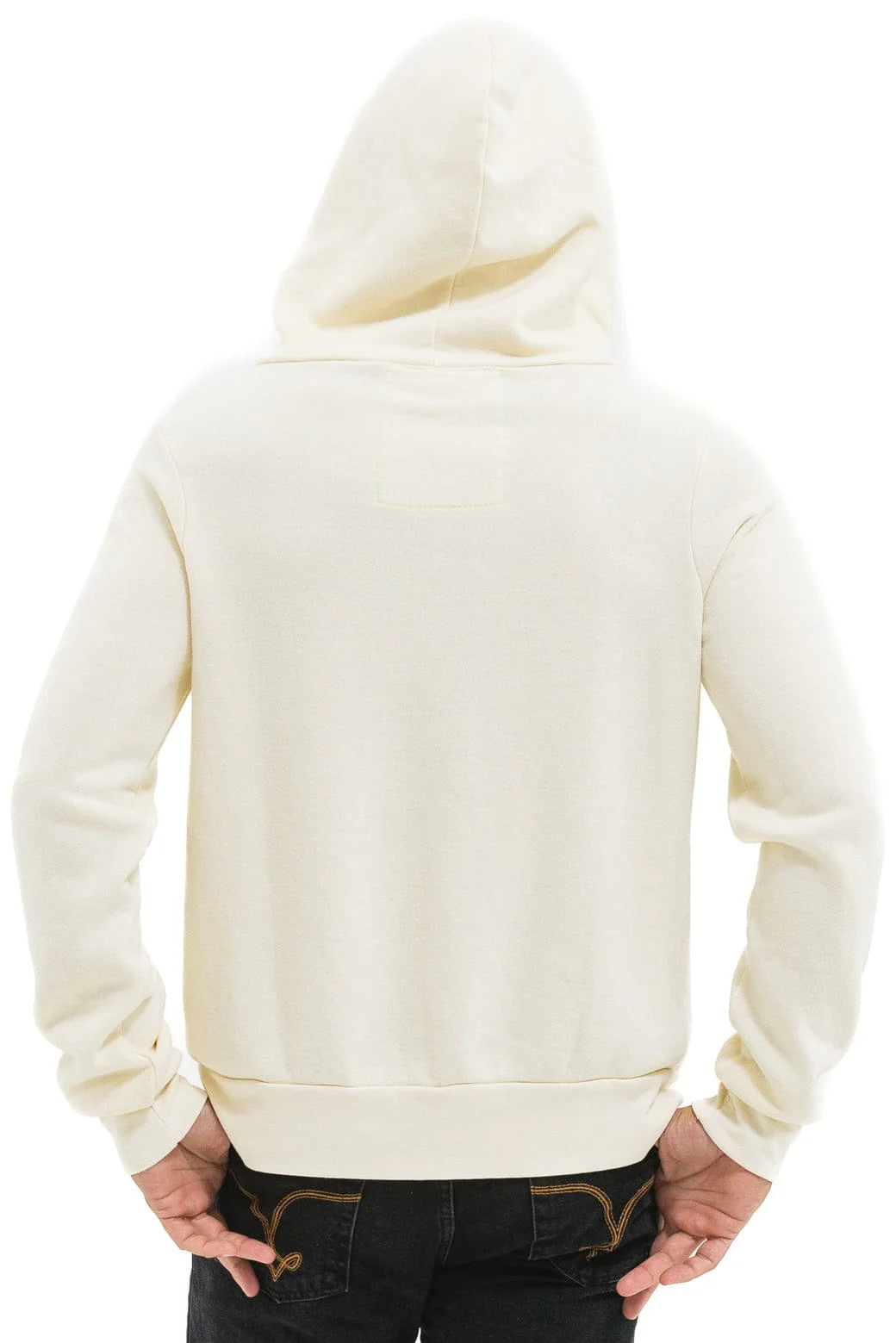 A person is seen from the back wearing an Aviator Nation 5 Stripe zip hoodie in cream, hood up, paired with dark jeans and hands on hips against a plain white background.