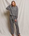 In front of a light-draped background, a person smiles in their relaxed fit hoodie and Collins Straight Sweatpant by Perfectwhitetee, featuring a white drawstring. Their hand is playfully raised by their head, with black flip-flops peeking from under the soft cotton fleece ensemble.
.