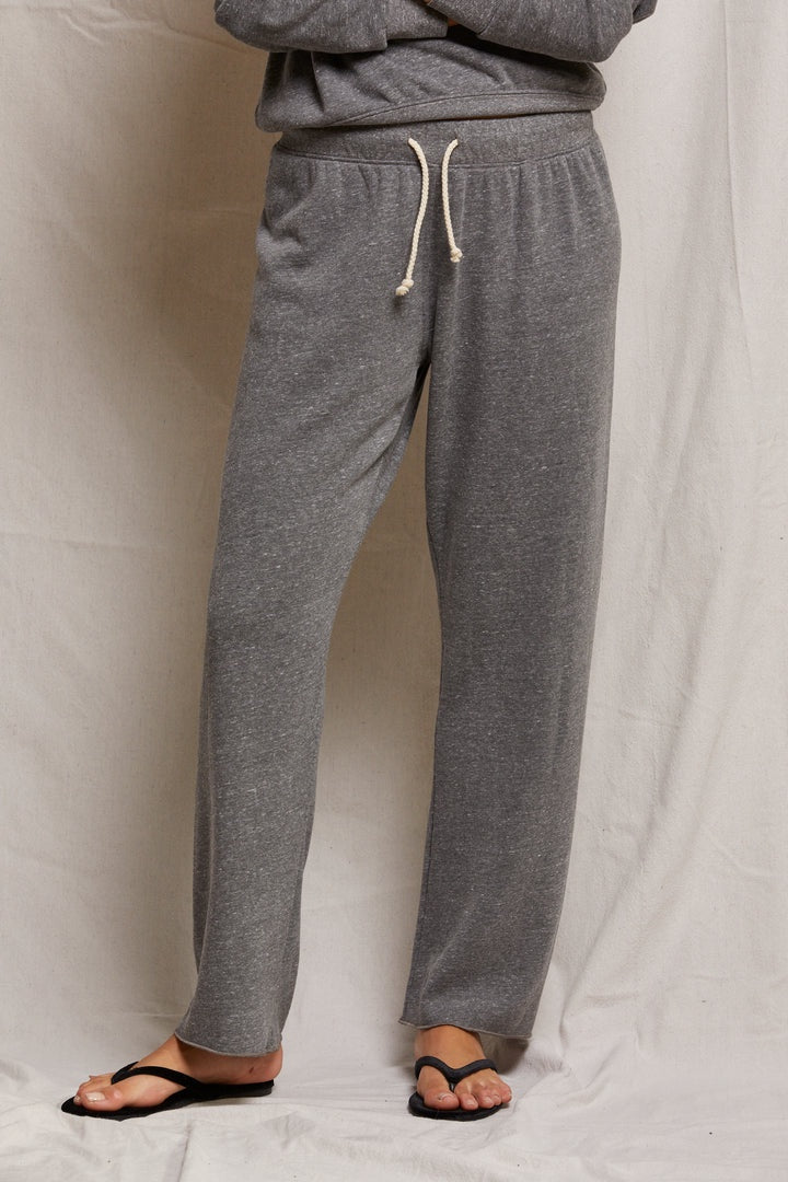 Standing against a light fabric background, only the lower half of a person is shown wearing Perfectwhitetee's Collins Straight Sweatpant in gray. This relaxed-fit pant features a white drawstring and pairs with a matching gray top and black sandals, highlighting its casual elegance.