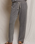 Standing against a light fabric background, only the lower half of a person is shown wearing Perfectwhitetee's Collins Straight Sweatpant in gray. This relaxed-fit pant features a white drawstring and pairs with a matching gray top and black sandals, highlighting its casual elegance.