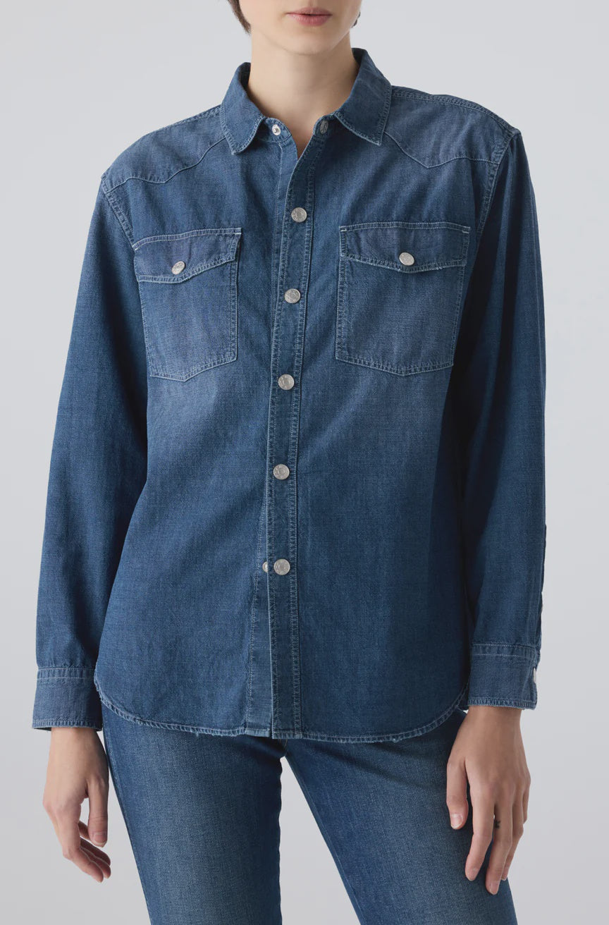 A person wears the AMO Denim Angela Shirt, a relaxed fit chambray top with front buttons and two chest pockets, paired with blue jeans against a plain light background.