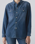 A person wears the AMO Denim Angela Shirt, a relaxed fit chambray top with front buttons and two chest pockets, paired with blue jeans against a plain light background.