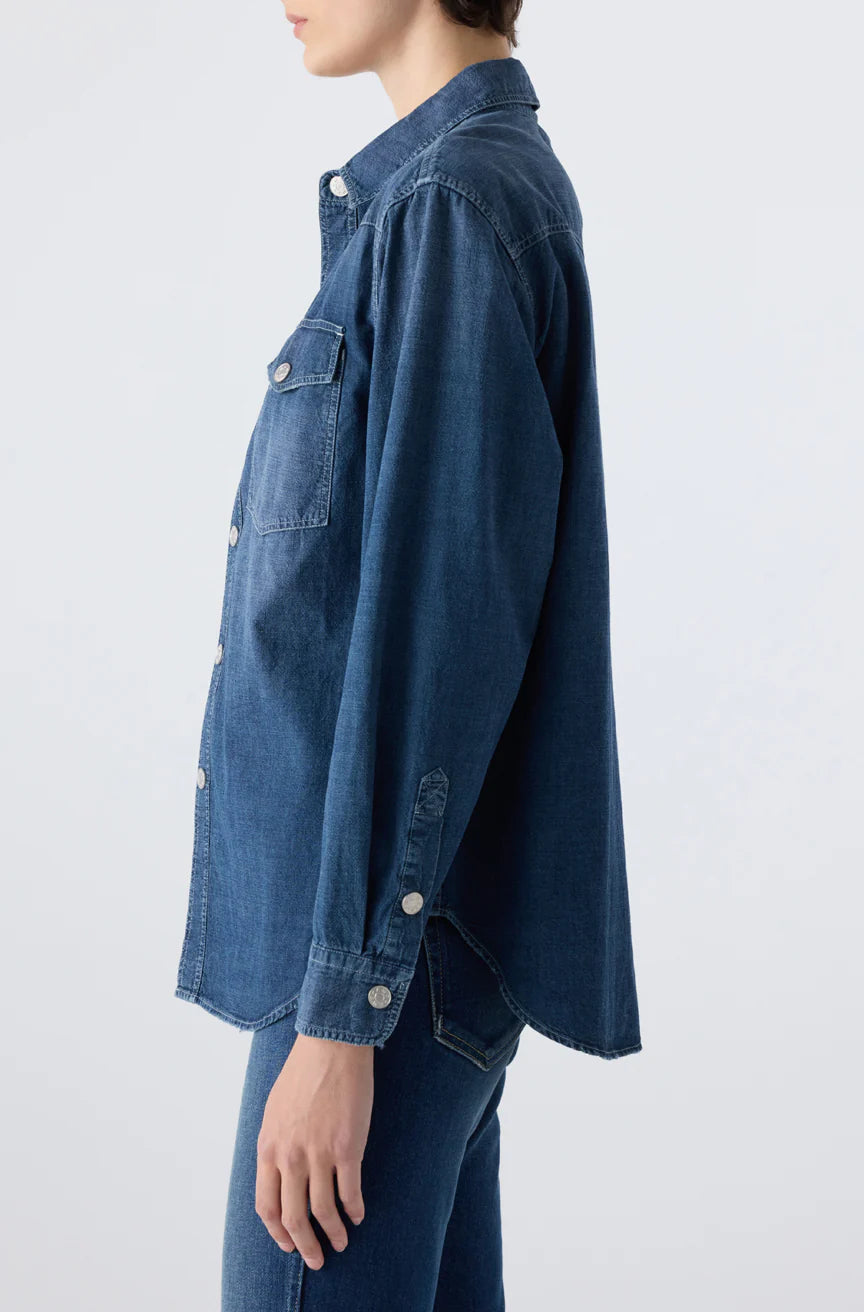 A person shown from the side wears the AMO Denim Angela Shirt—a relaxed fit, long sleeve blue denim button-up with front pockets—paired with matching jeans against a plain background.