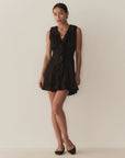 A woman stands against a plain light wall wearing the DOEN Henrietta Dress, a sleeveless black mini dress with lace-trimmed ruffles at the neckline and hem. She pairs it with black flats and gazes to the side in a relaxed pose.