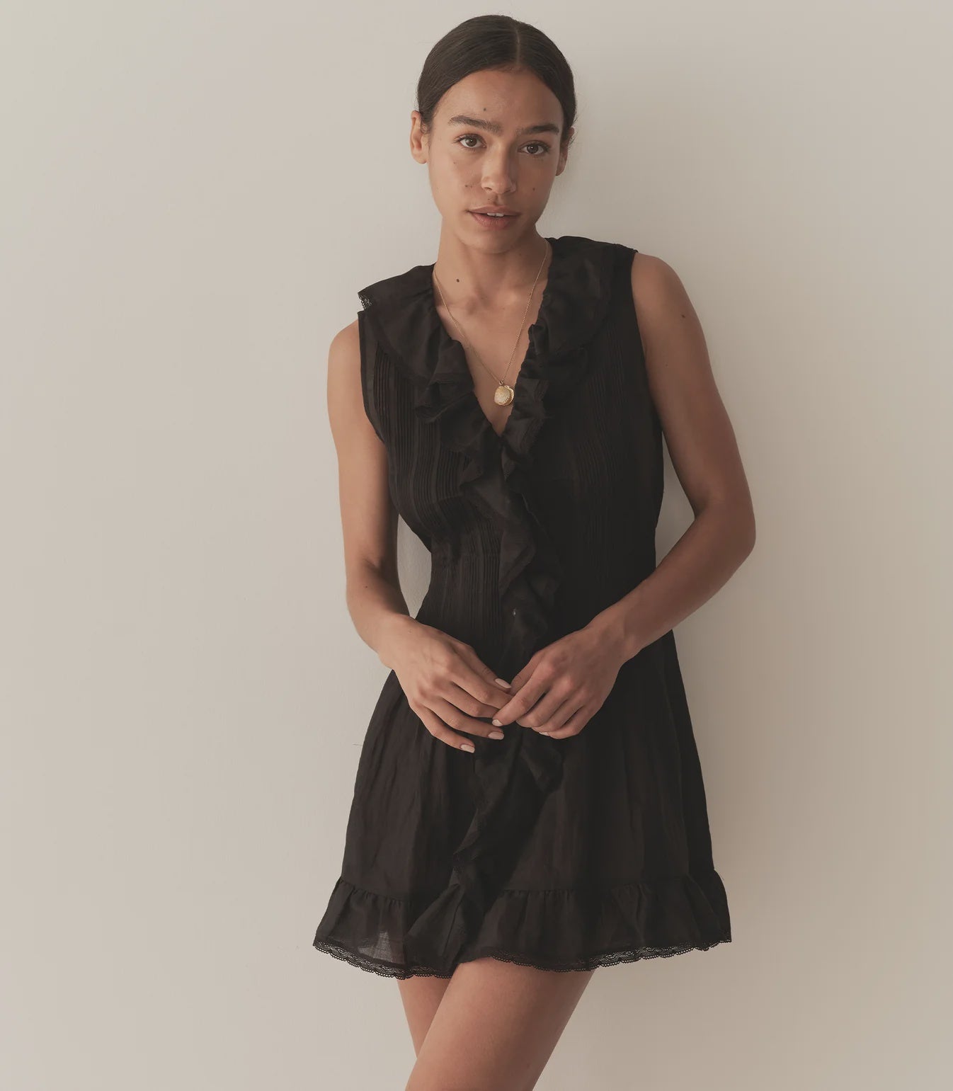 A woman with dark hair pulled back wears the DOEN Henrietta Dress, a sleeveless mini with lace-trimmed ruffle details at the neckline and hem, standing against a plain light-colored wall.