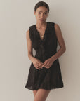 A woman with dark hair pulled back wears the DOEN Henrietta Dress, a sleeveless mini with lace-trimmed ruffle details at the neckline and hem, standing against a plain light-colored wall.