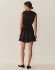 A woman stands facing away against a light wall, wearing DOEN's Henrietta Dress—a sleeveless mini dress with a lace-trimmed ruffle hem and collar, waist tie bow—and flat black shoes. Her hair is tied back.