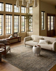 A cozy living room in a Scottsdale bungalow, featuring a large beige sofa, a white round coffee table, and a wicker chair. The room includes large windows, wooden floors, and beige walls decorated with touches of greenery. The room is anchored by a Sage / Navy rug from Loloi Rugs.