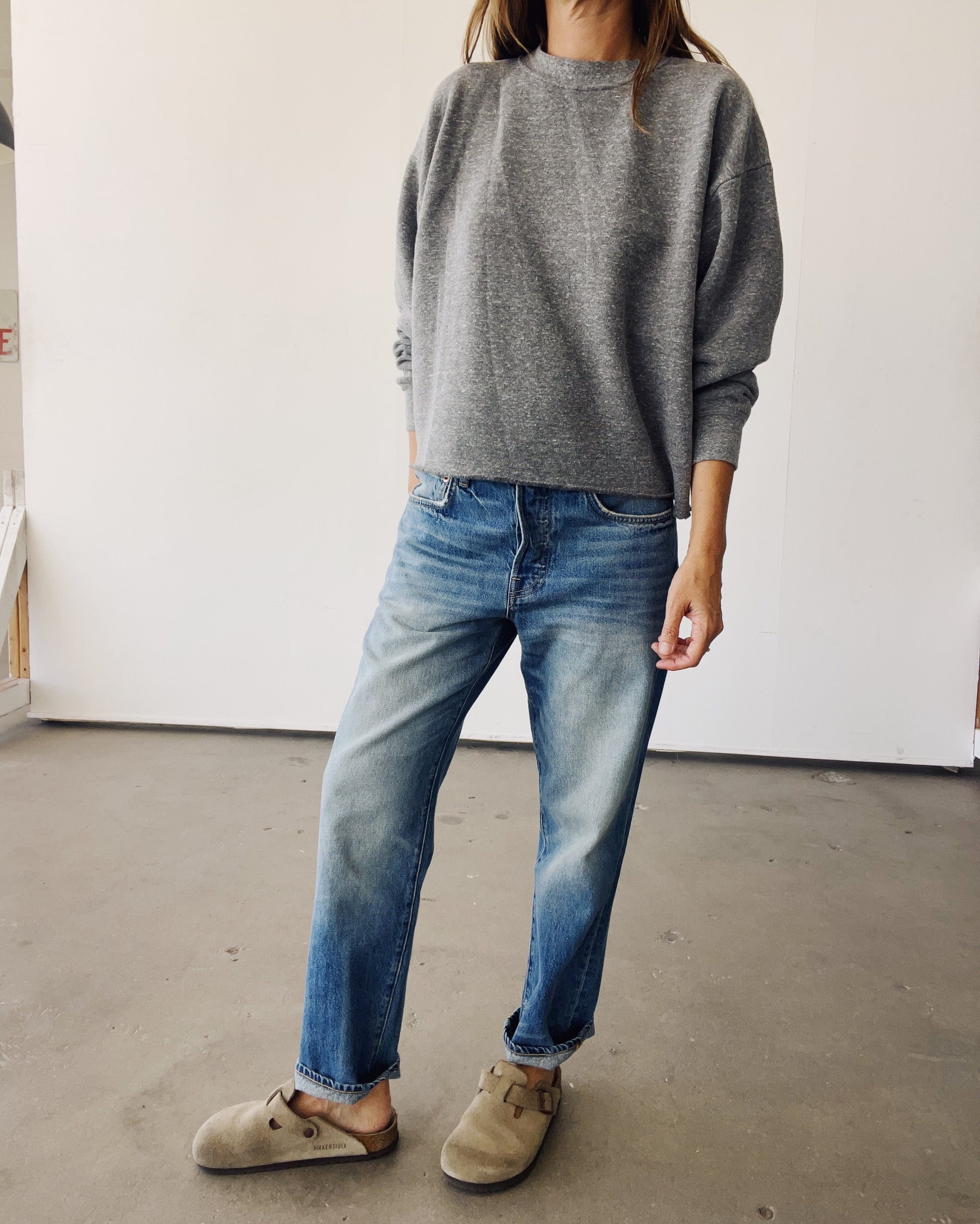 Indoors, a person wears Hey Gang's The Boxy Crop Sweatshirt in gray, blue jeans, and tan slip-on shoes. Their right hand rests in a pocket; their head is out of frame.