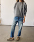 Indoors, a person wears Hey Gang's The Boxy Crop Sweatshirt in gray, blue jeans, and tan slip-on shoes. Their right hand rests in a pocket; their head is out of frame.