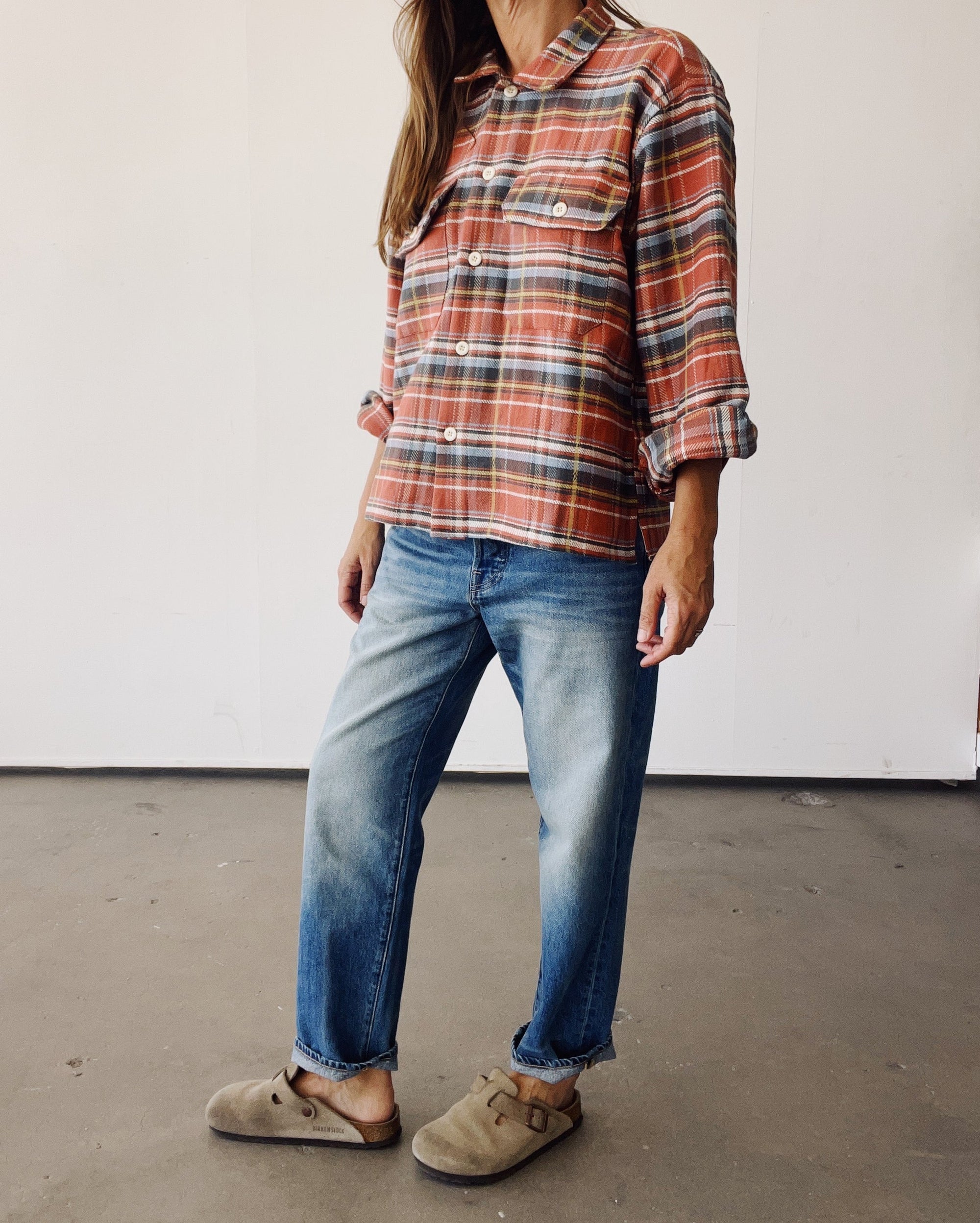 A person stands indoors in a plain, light setting, wearing Hey Gang's The Work Shirt in rust plaid with a boxy fit, paired with faded blue jeans and tan slip-on shoes. Their head is partially out of view.