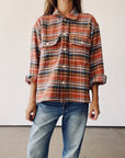 A person wears The Work Shirt by Hey Gang—a rust plaid, boxy-fit shirt with rolled-up sleeves—paired with blue jeans, standing against a plain white background.