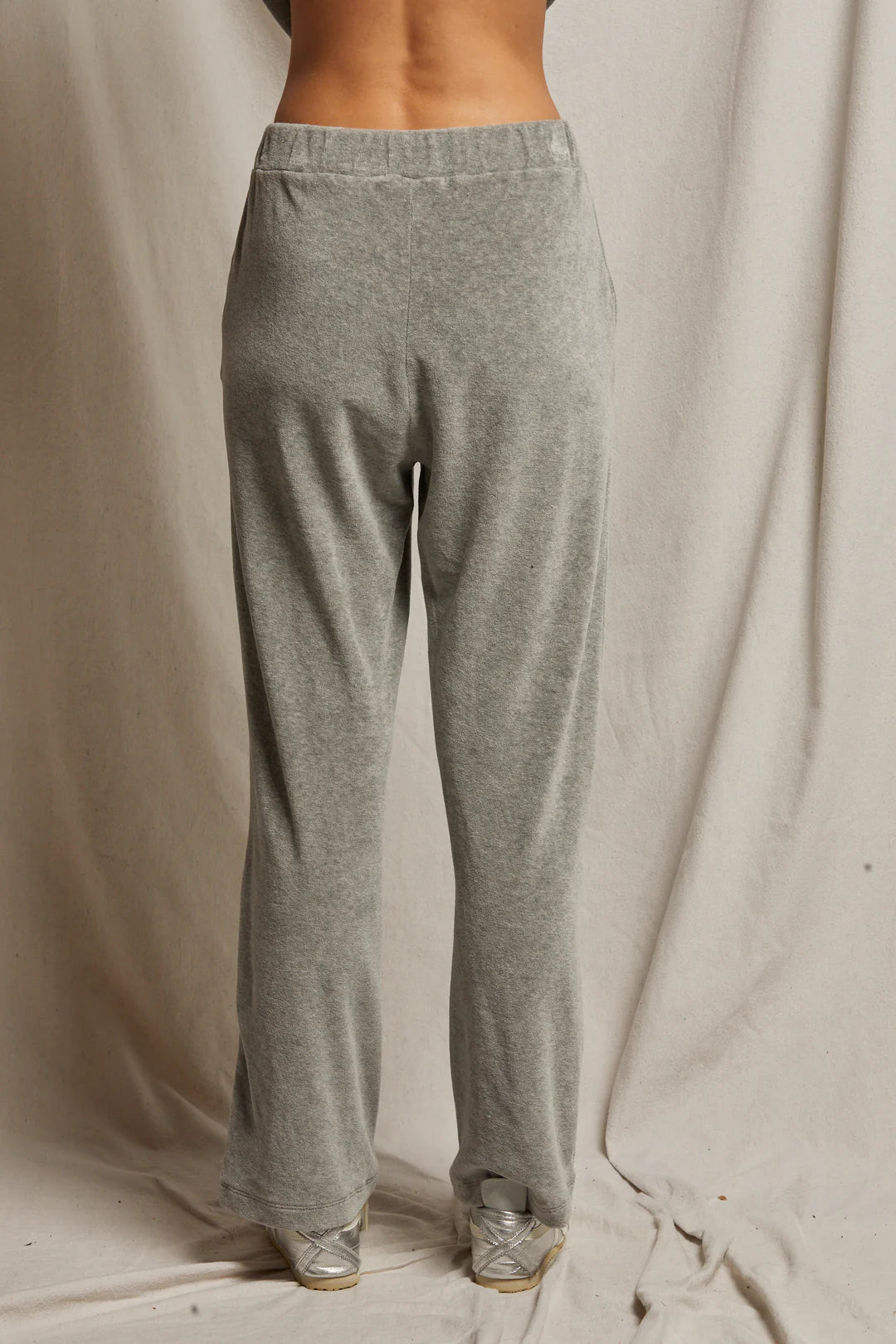 A person wearing the Holly Velour Wide Leg Sweatpant by Perfectwhitetee stands with their back to the camera. The grey sweatpants have a loose fit, and they are on a fabric-draped floor, with grey sneakers on their feet.