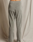 A person wearing the Holly Velour Wide Leg Sweatpant by Perfectwhitetee stands with their back to the camera. The grey sweatpants have a loose fit, and they are on a fabric-draped floor, with grey sneakers on their feet.