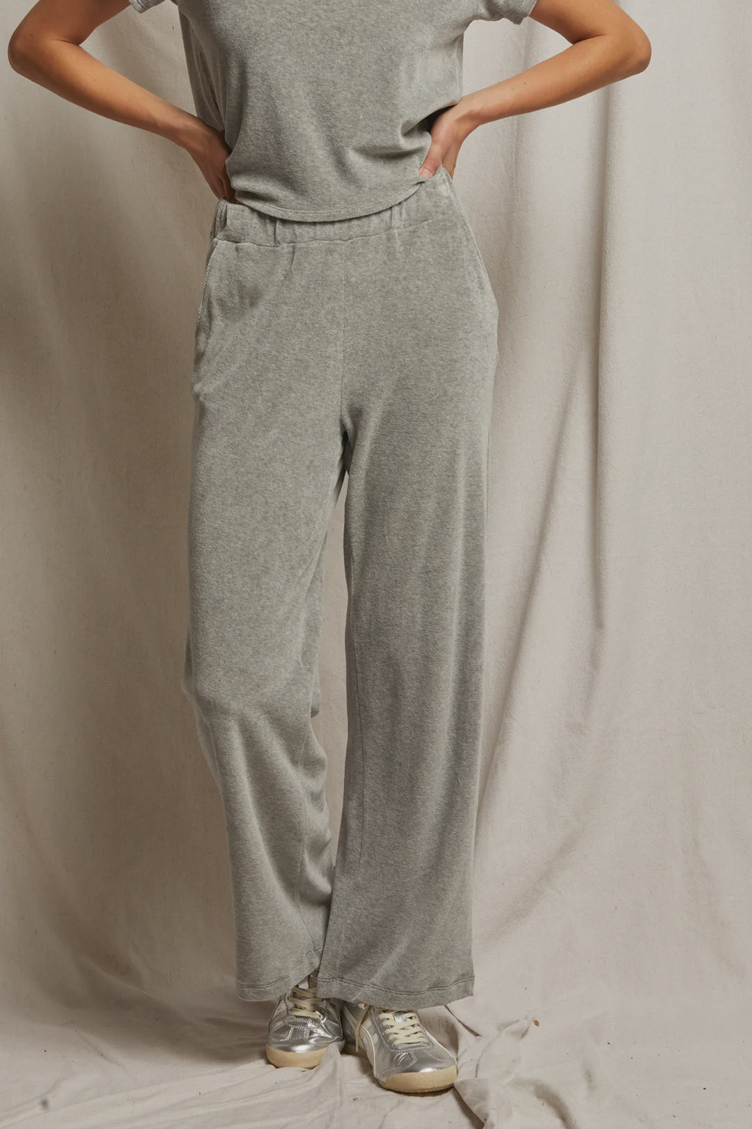 A person stands against a beige backdrop wearing a light gray t-shirt and the Holly Velour Wide Leg Sweatpant from Perfectwhitetee, with their hands tucked into the pockets and metallic sneakers on their feet.