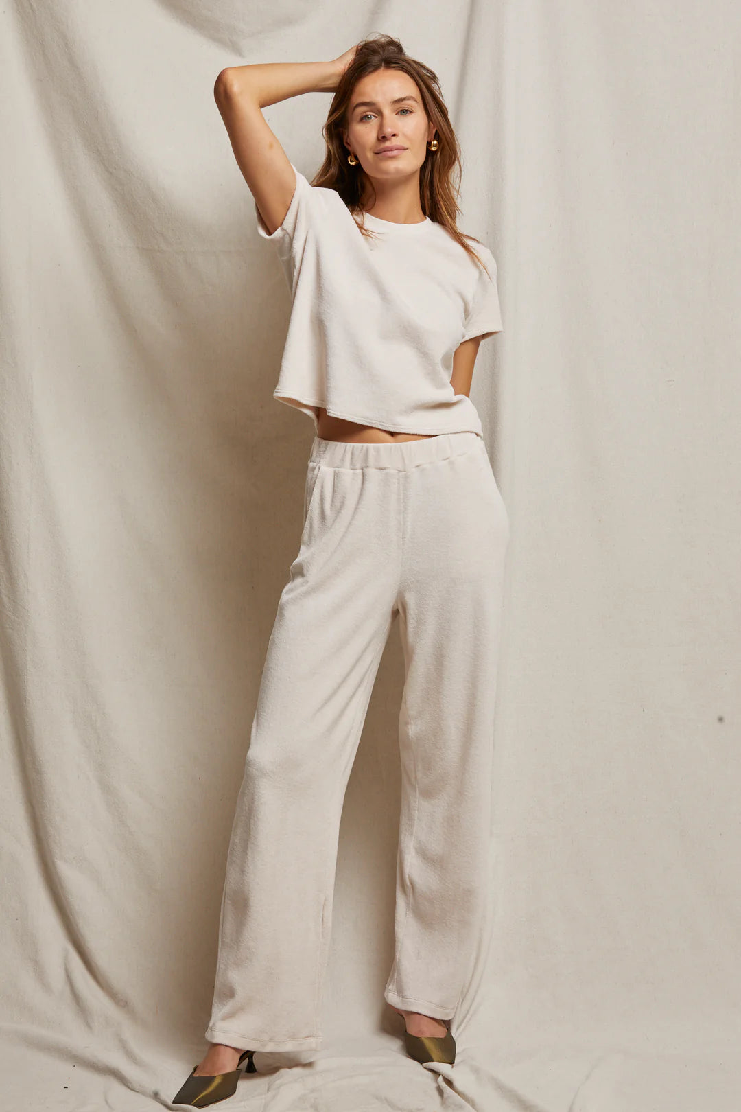 Against a neutral fabric backdrop, an individual is dressed in a casual light beige ensemble, featuring the Holly Velour Wide Leg Sweatpant from Perfectwhitetee and a short-sleeve top. With one hand resting on their head, they sport plush velour pointed shoes and offer a slight smile to the camera.