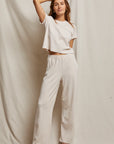 Against a neutral fabric backdrop, an individual is dressed in a casual light beige ensemble, featuring the Holly Velour Wide Leg Sweatpant from Perfectwhitetee and a short-sleeve top. With one hand resting on their head, they sport plush velour pointed shoes and offer a slight smile to the camera.