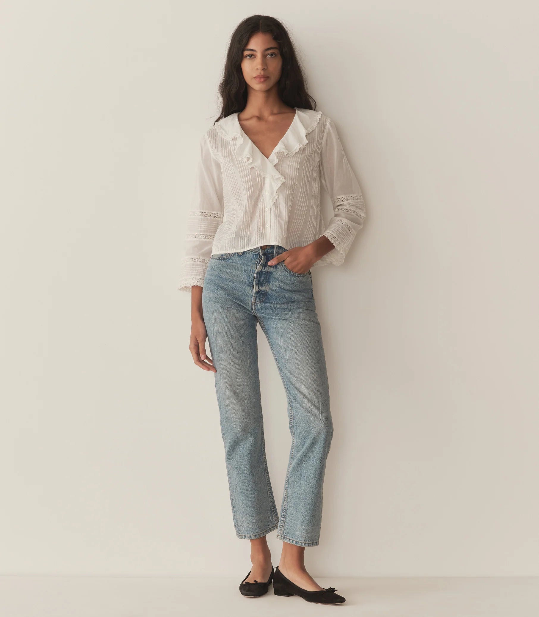 A woman stands against a plain light-colored wall, wearing the DOEN Huguette Top, light blue high-waisted jeans, and black ballet flats. She has long dark hair and a relaxed, confident stance with one hand in her pocket.