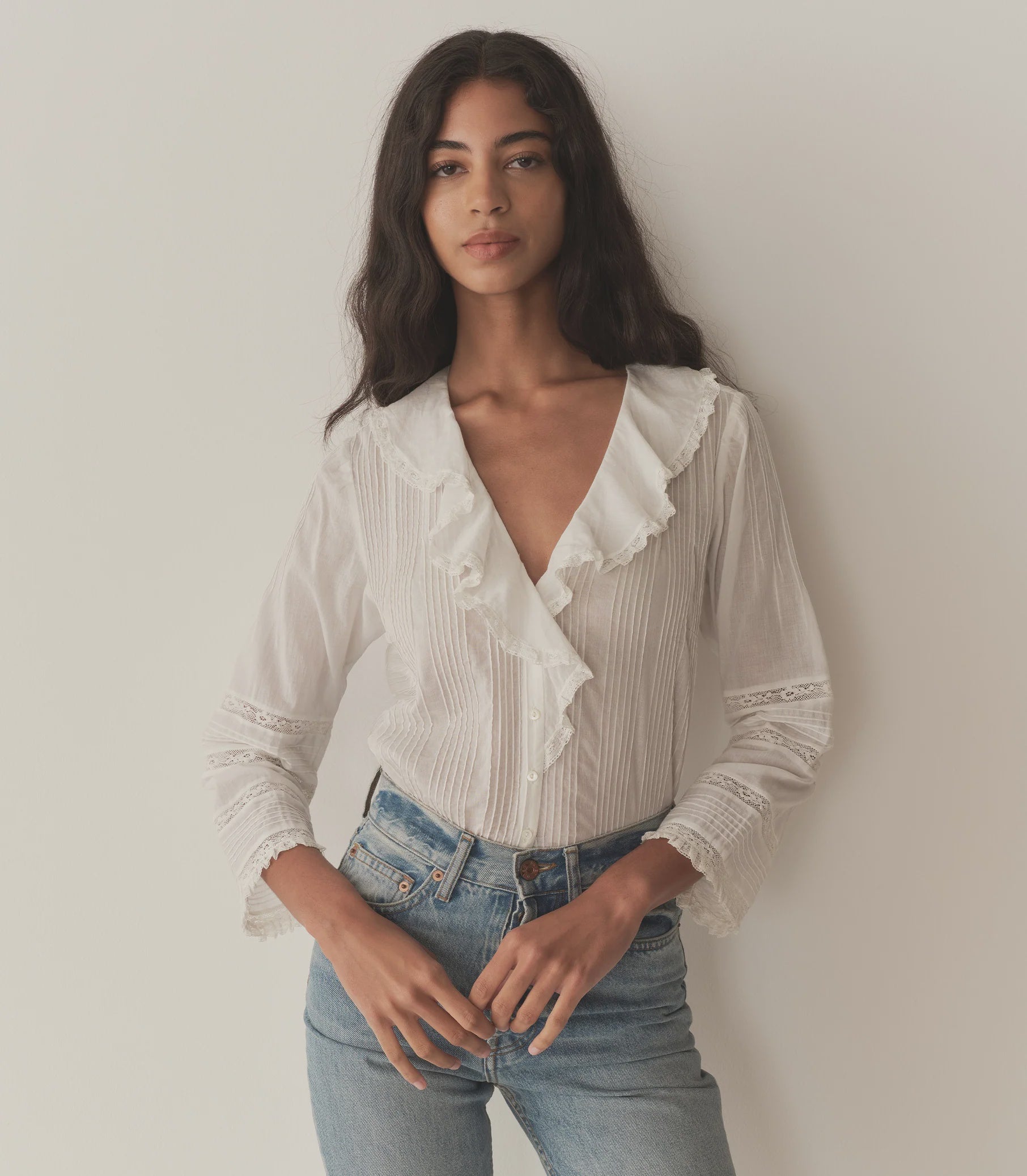 A woman with long dark hair wears the stylish DOEN Huguette Top and blue jeans, standing against a plain light wall and looking at the camera with a neutral expression.