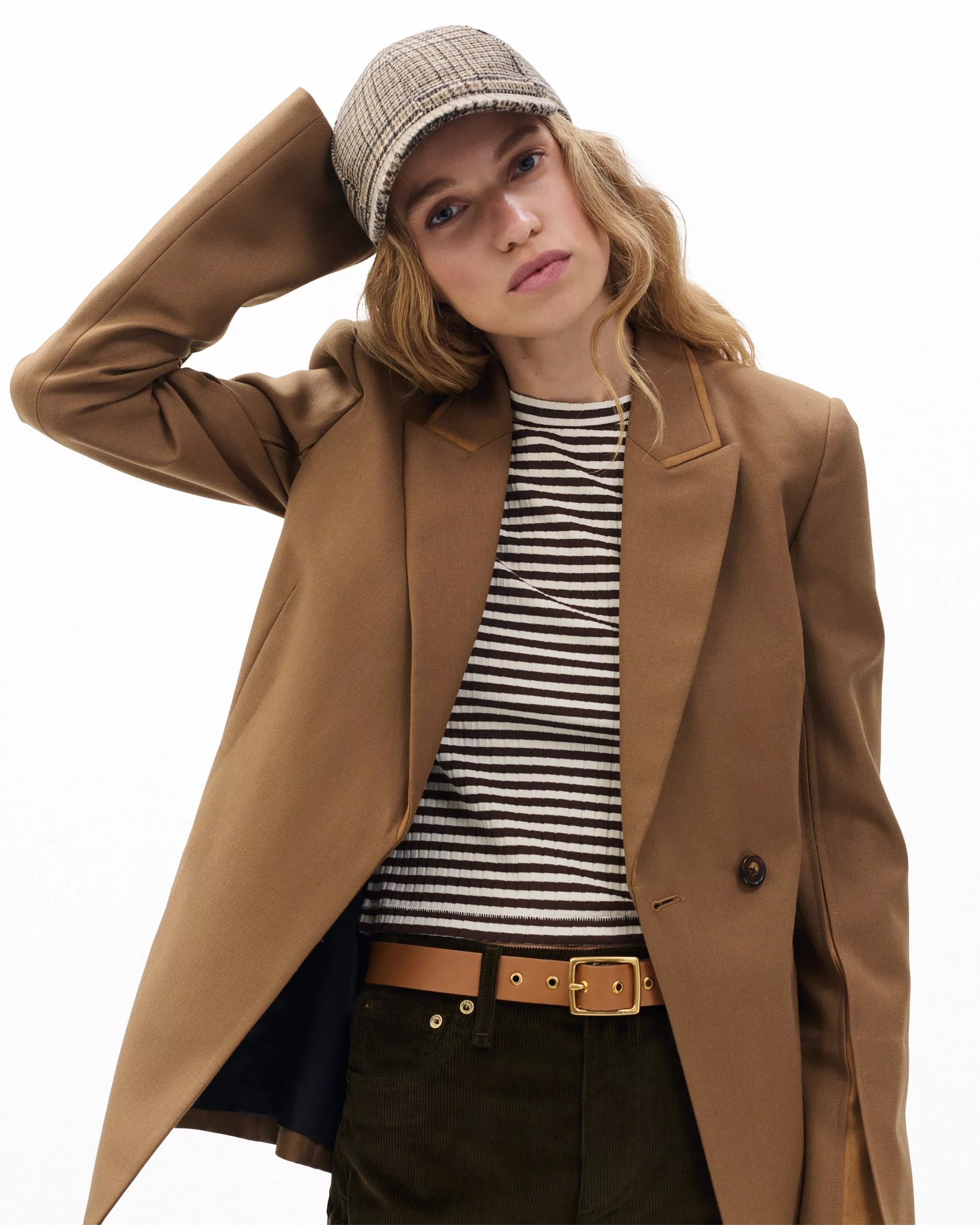 A woman wears a Rag & Bone harlow baseball cap, brown blazer, black and white striped shirt, olive pants, and a tan belt, standing with one hand behind her head against a plain white background.