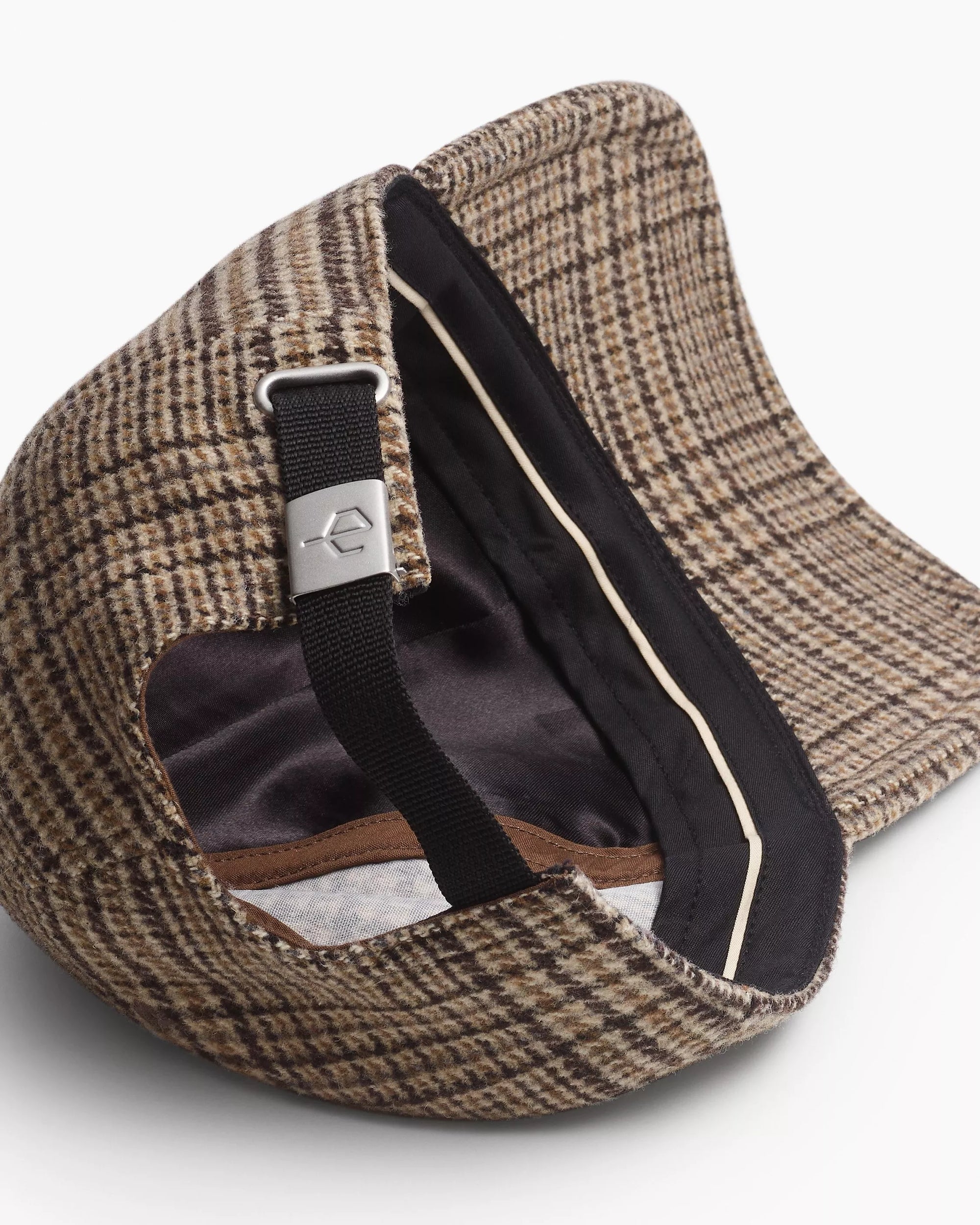 A close-up of the Rag & Bone harlow baseball cap showcases its brown plaid pattern, black adjustable strap with a metal clasp, and intricate stitching along the inner brim.