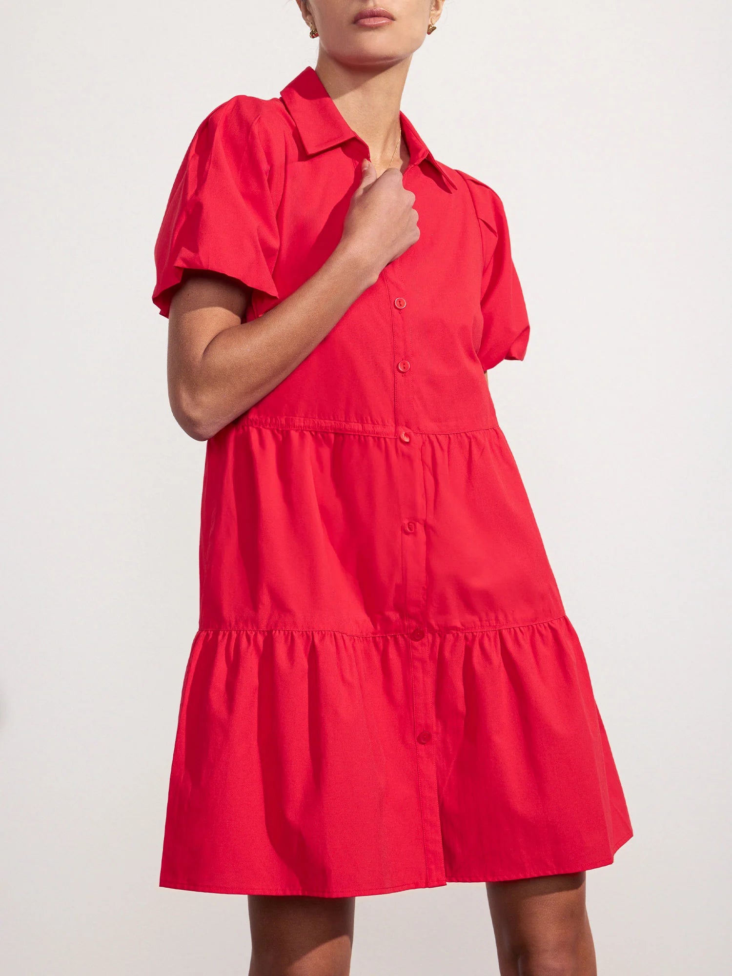 A person wears the bright red BROCHU WALKER HAVANA MINI DRESS, a short-sleeve, button-down mini dress with a tiered skirt and collar, standing against a plain light background.
