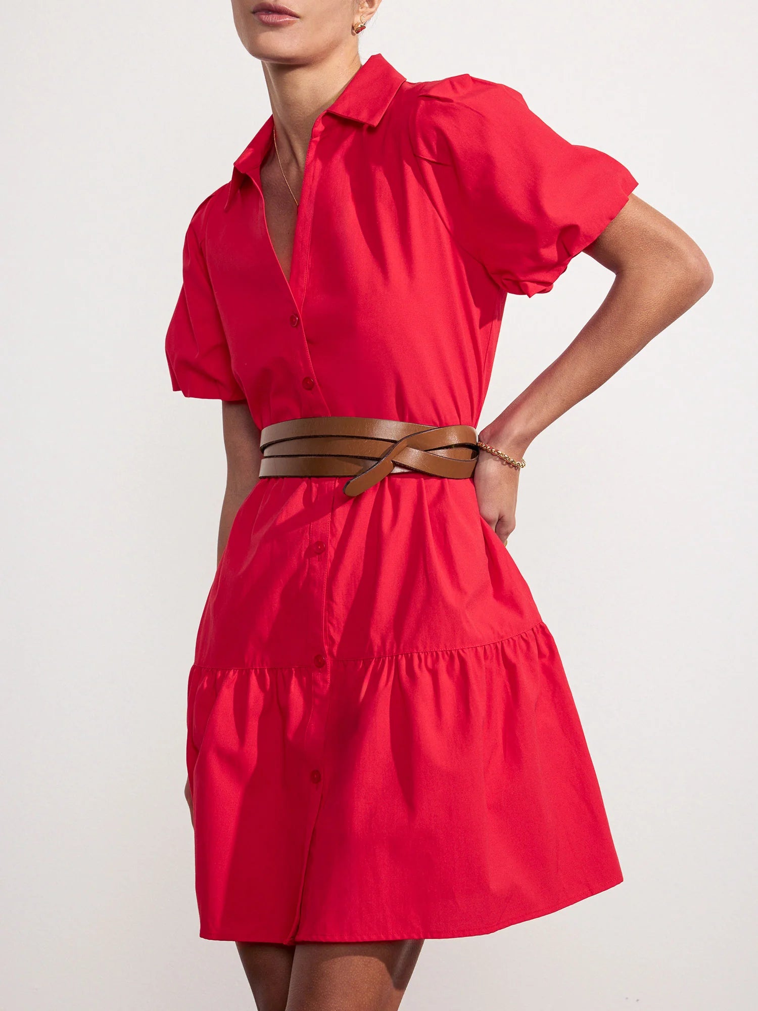 A woman wearing the BROCHU WALKER HAVANA MINI DRESS in bright red, featuring puff sleeves and a tiered skirt with a brown wrap belt, poses against a plain white background. Her head is slightly tilted and her face is partially cropped.