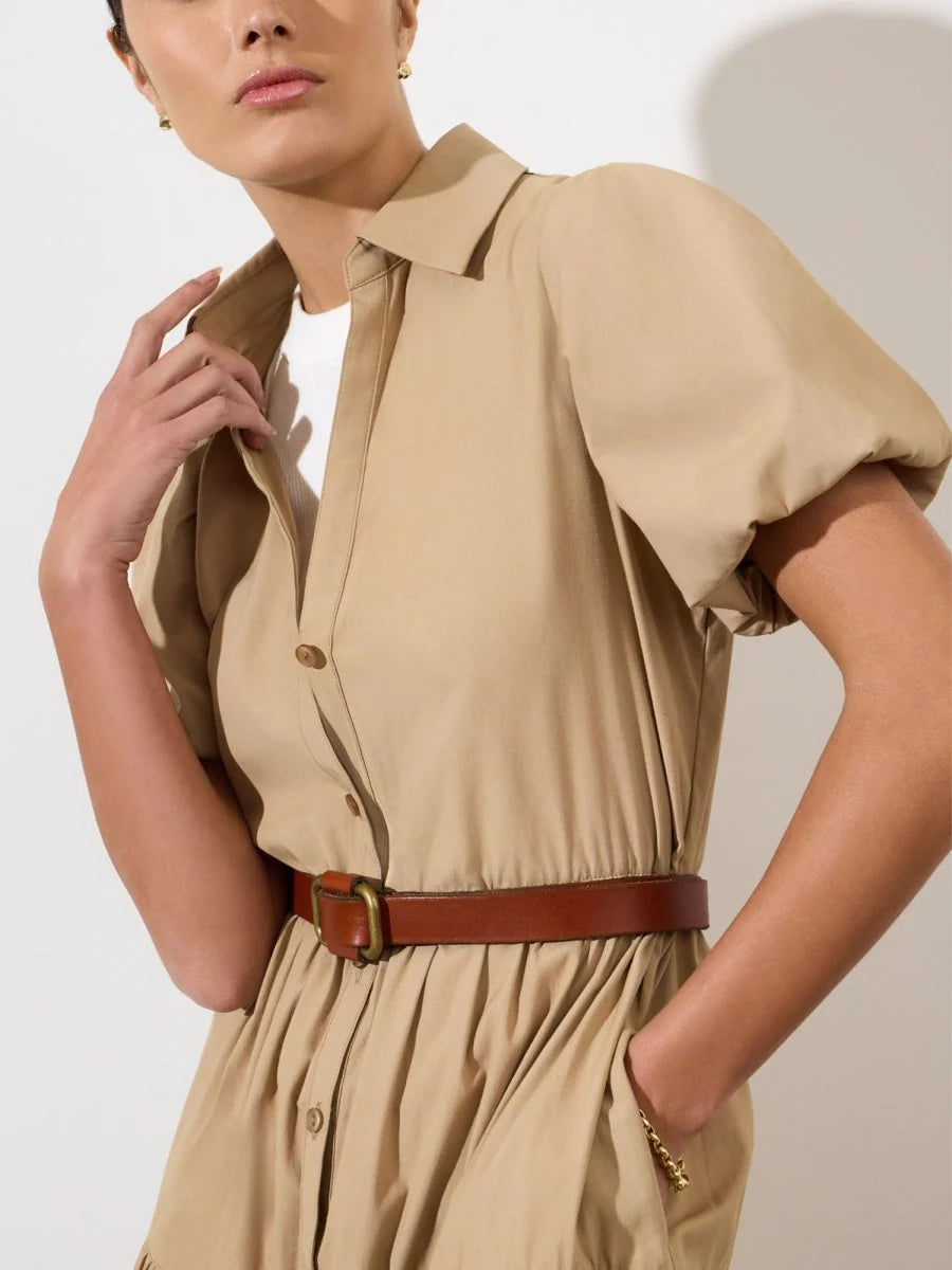 A person wearing the BROCHU WALKER HAVANA DRESS, a beige puff sleeve style paired with a brown belt and white top underneath, stands against a plain light background.