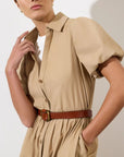 A person wearing the BROCHU WALKER HAVANA DRESS, a beige puff sleeve style paired with a brown belt and white top underneath, stands against a plain light background.
