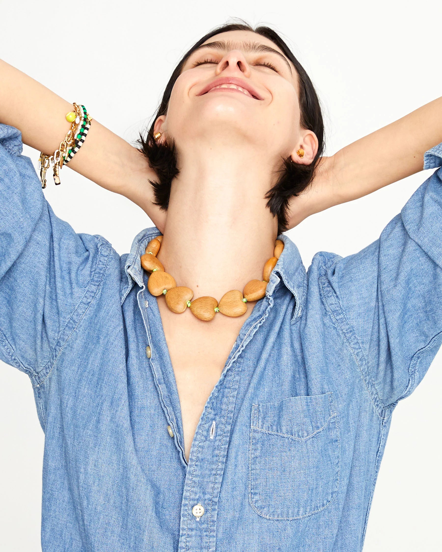 A person wearing a light denim button-up shirt and the Clare Vivier Shortie Strap/Heart, a necklace with wooden beads and neon yellow thread, smiles with eyes closed and arms raised behind their head against a plain white background.