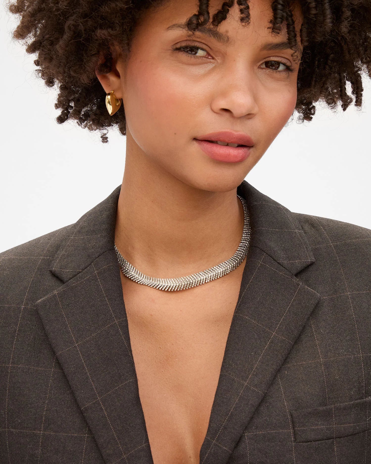 A woman with curly hair wears a dark plaid blazer, gold hoop earrings, and the Clare Vivier Herringbone Collar necklace. She gazes slightly off-camera with a soft expression against a plain background.