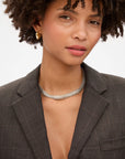 A woman with curly hair wears a dark plaid blazer, gold hoop earrings, and the Clare Vivier Herringbone Collar necklace. She gazes slightly off-camera with a soft expression against a plain background.