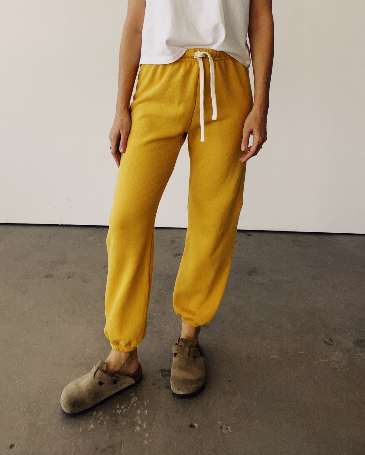 Hey Gang Goldenrod sweatpants and white tee styled casually with brown slippers on a modern concrete floor.