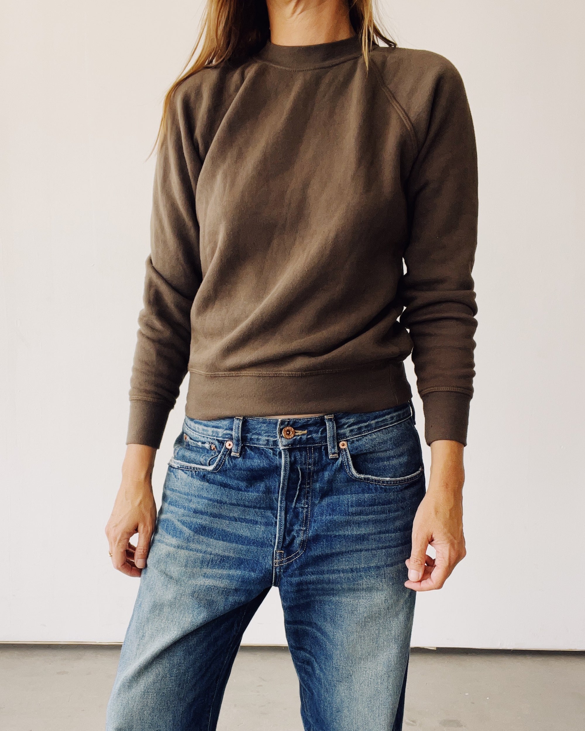 A person wears The Sweatshirt by Hey Gang in brown with blue jeans, standing against a plain light background; their face is partially out of frame.