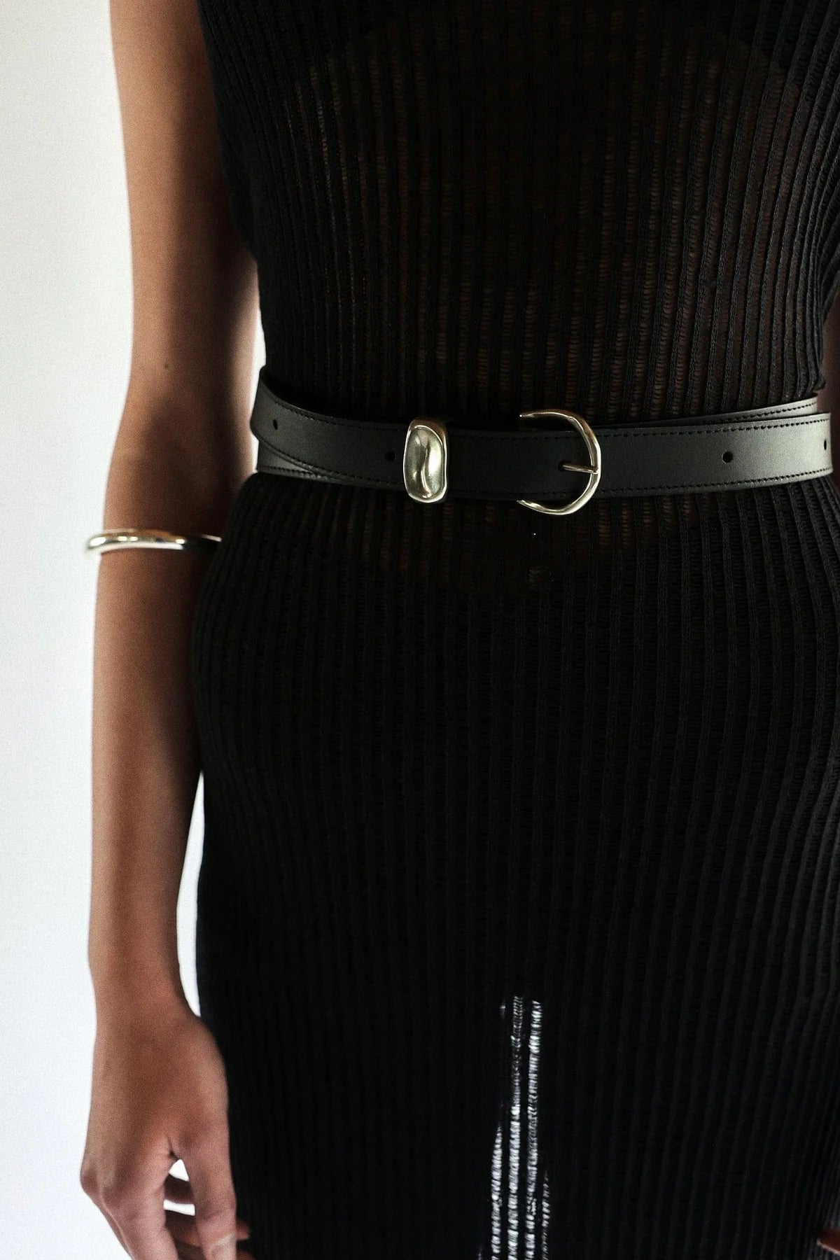 Dressed in a black ribbed dress, a person wears a Janessa Leone Myra DW Belt with artisan-crafted silver hardware. Their left arm is adorned with a silver bangle, all set against a plain white background.