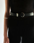 Dressed in a black ribbed dress, a person wears a Janessa Leone Myra DW Belt with artisan-crafted silver hardware. Their left arm is adorned with a silver bangle, all set against a plain white background.