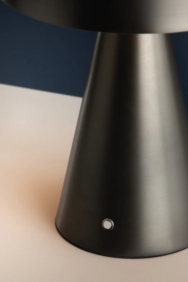 A close-up of the Hudson Valley Hopkins Rechargeable lamp base, featuring a modern metallic cone shape with a small round button, set against a contrasting dark and light background.
