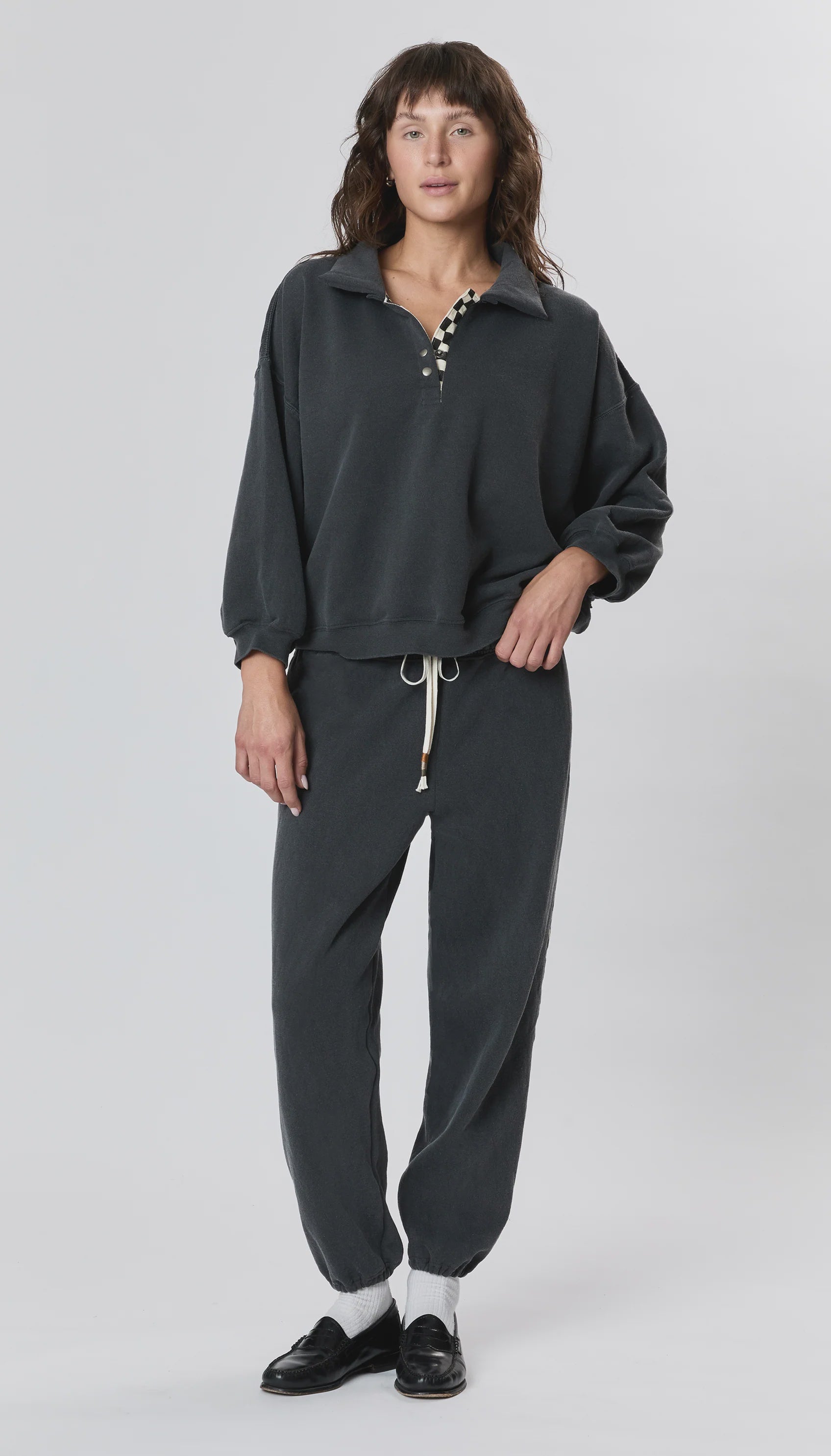 A person stands facing forward in a loose dark gray pullover and Ragabond Studio’s Studio Sweats Checkerboard jogger pants. They have medium-length brown hair, black shoes with white socks, and a plain light background.