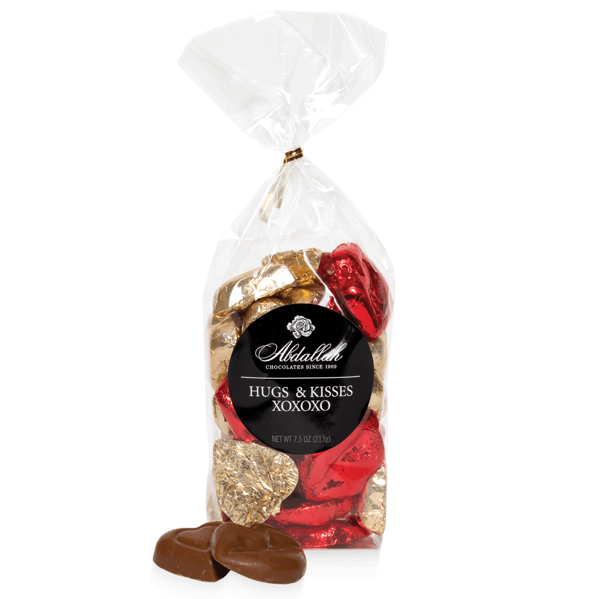 A clear 7.5 oz bag from Abdallah reveals heart-shaped chocolates wrapped in red and gold foil, showcasing their pure milk chocolate delight. The label reads "Milk Chocolate Bags" and "Hugs & Kisses XOXOXO." Three unwrapped chocolates are displayed in front.