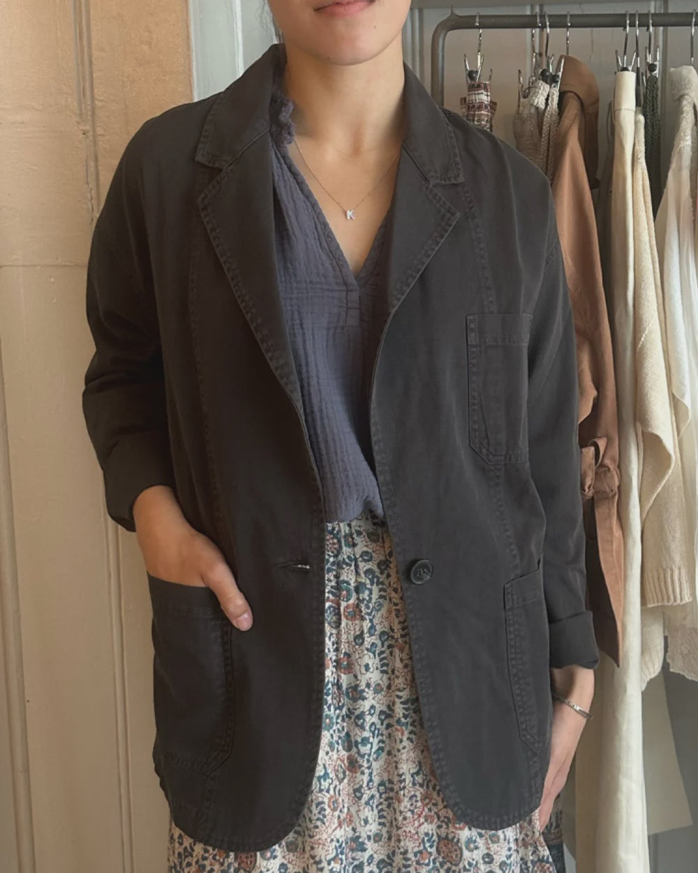 A person stands indoors wearing the Jonnie Jacket by Xirena over a blue blouse and floral skirt. One hand is in their pocket, and clothes hang on a rack behind them. Their face is partially out of frame.