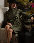 A woman in THE SHIRT's Wrap Front Dress in dark green and a black cowboy hat sits on a beige chair indoors, one leg crossed over the other, surrounded by glassware and decorative items.
