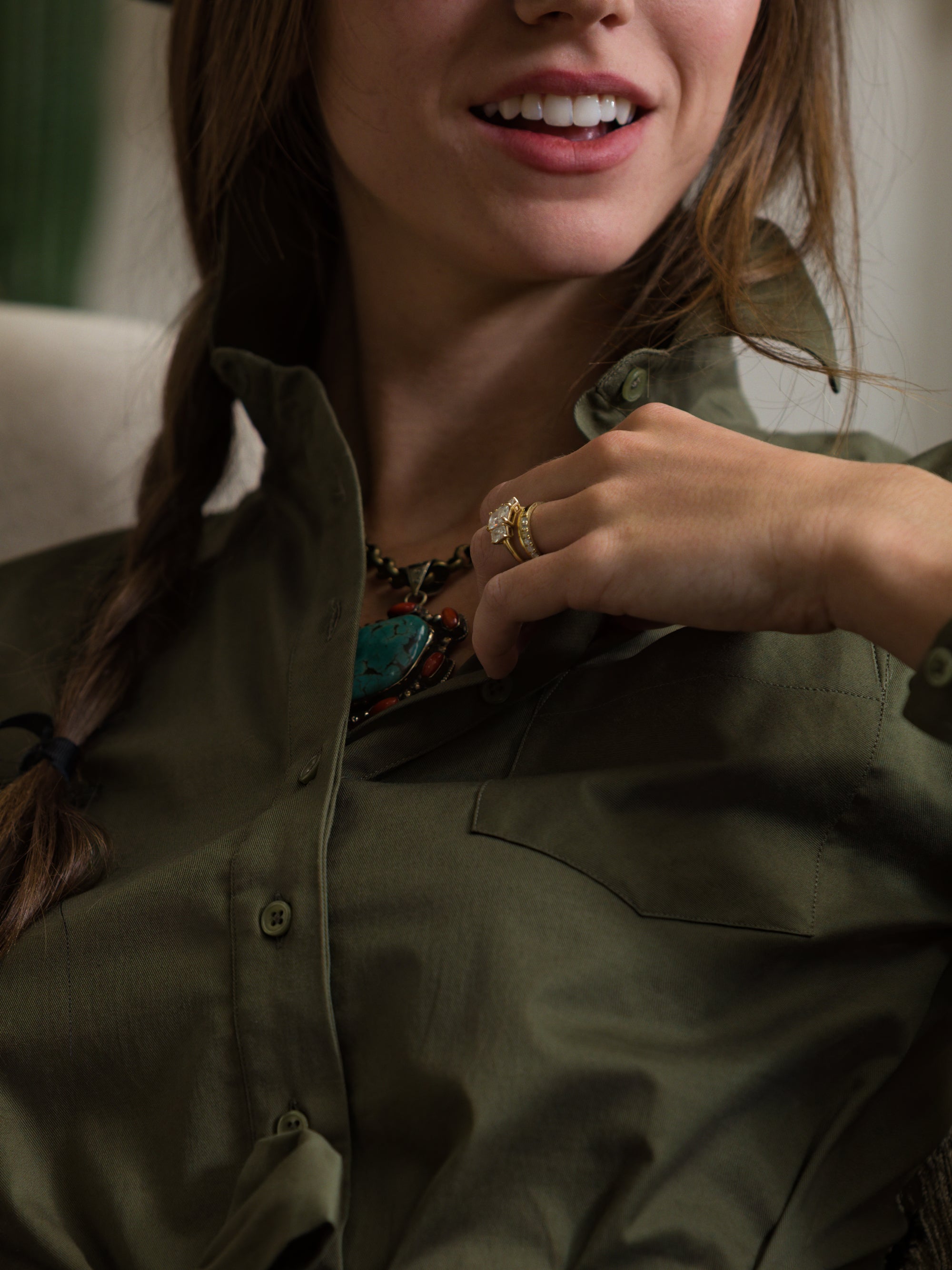 A woman in THE SHIRT’s olive-green The Wrap Front Dress smiles slightly, revealing part of her teeth. She has a braid and wears a turquoise necklace and gold ring, with her hand resting near her collar.