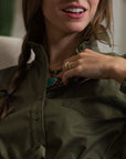 A woman in THE SHIRT’s olive-green The Wrap Front Dress smiles slightly, revealing part of her teeth. She has a braid and wears a turquoise necklace and gold ring, with her hand resting near her collar.
