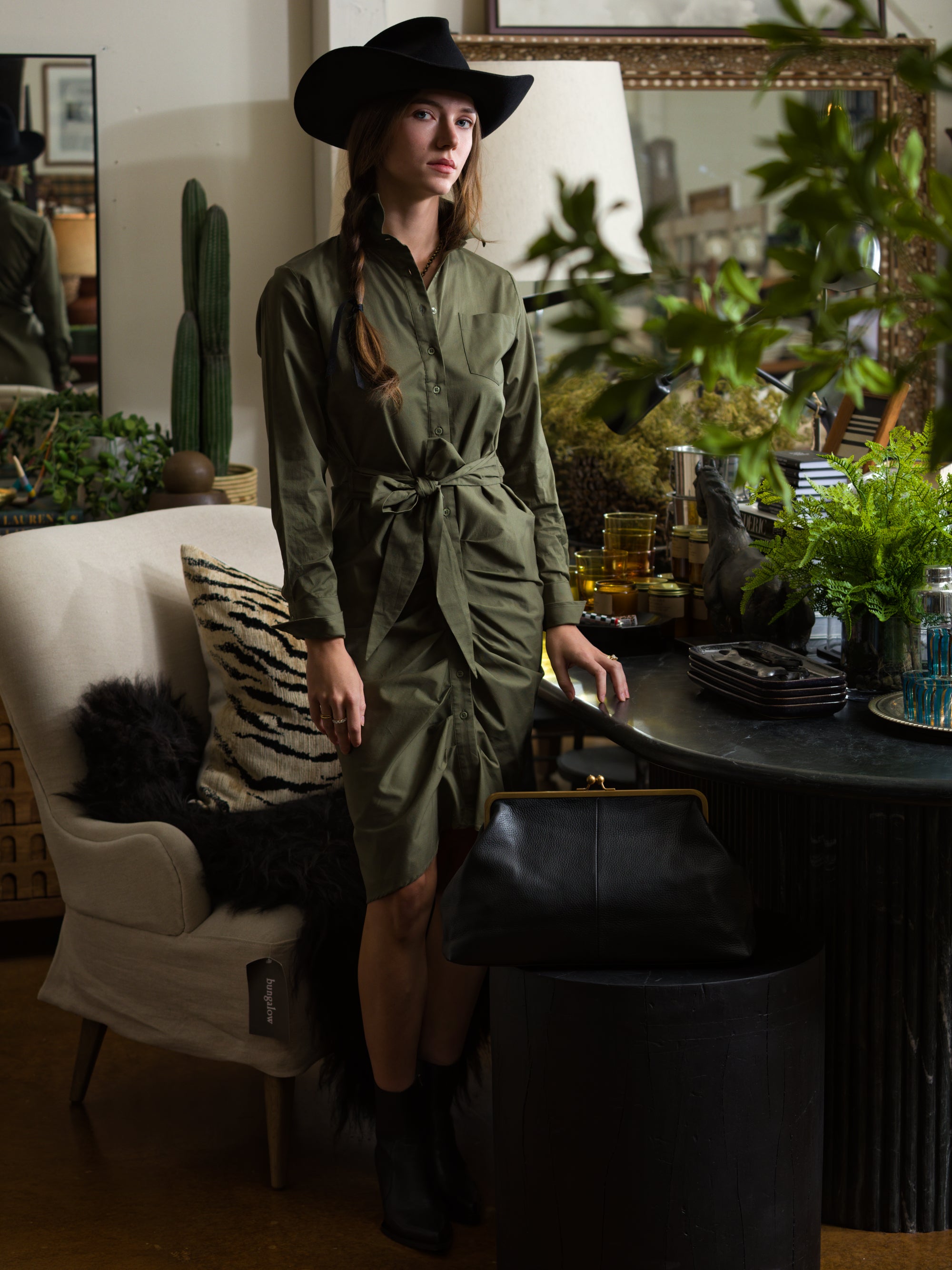 A woman wears The Wrap Front Dress by THE SHIRT in olive green, styled with a black hat. She stands indoors beside plants and glassware near a beige armchair and a large black bag on a round stool.
