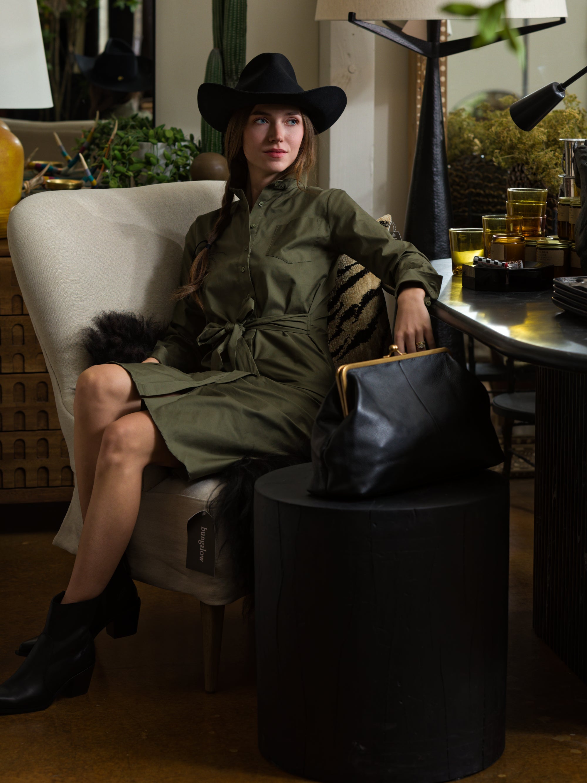 A woman wears THE SHIRT’s The Wrap Front Dress—an olive-green, long-sleeve wrap dress—styled with a black hat and ankle boots, as she sits on a cream armchair. A black handbag rests on the nearby round table in the eclectic room.