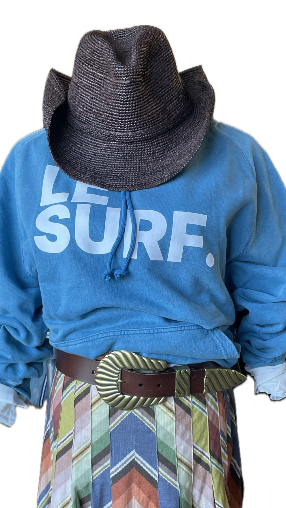 A headless mannequin wears a black woven hat, Ping Pong Surf Club's Le Surf Hoodie with "LE SURF." text, a colorful patterned skirt, and a wide brown belt with a large metallic buckle. The background is white.